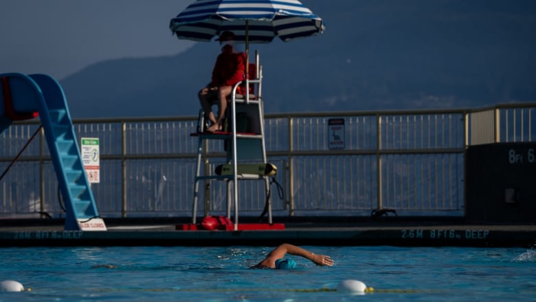 A public pool can be seen with a few people swimming and a lifeguard sitting close by.