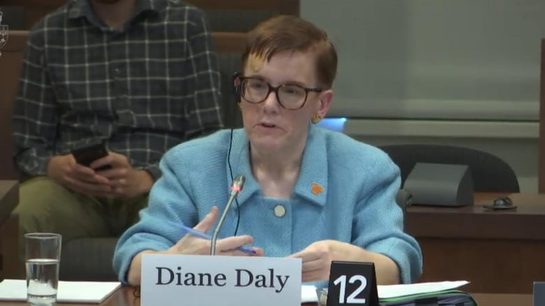 A woman in a blue button up sweater sits at a table and speaks into a microphone.