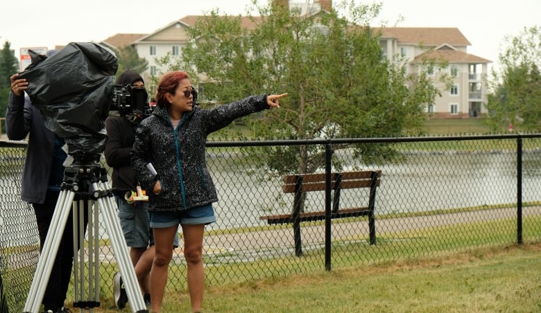 A woman in shorts and a black jacket is seen directing a man who is standing with a huge tripod and film camera.