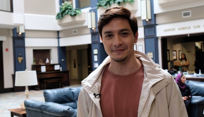 A close-up shot of a guy in a brown T-shirt. He is wearing a light jacket and is seen smiling.