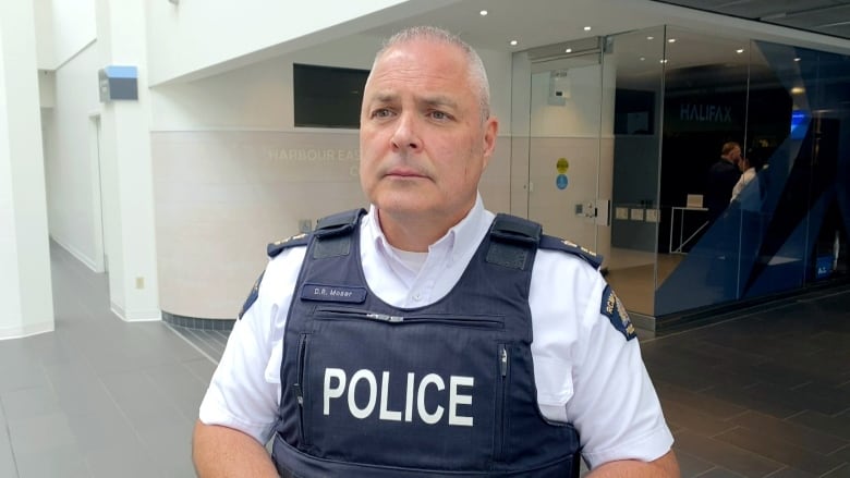 A white man in a police uniform and tactical vest with the word POLICE in white lettering stands in an atrium with a glass door behind him