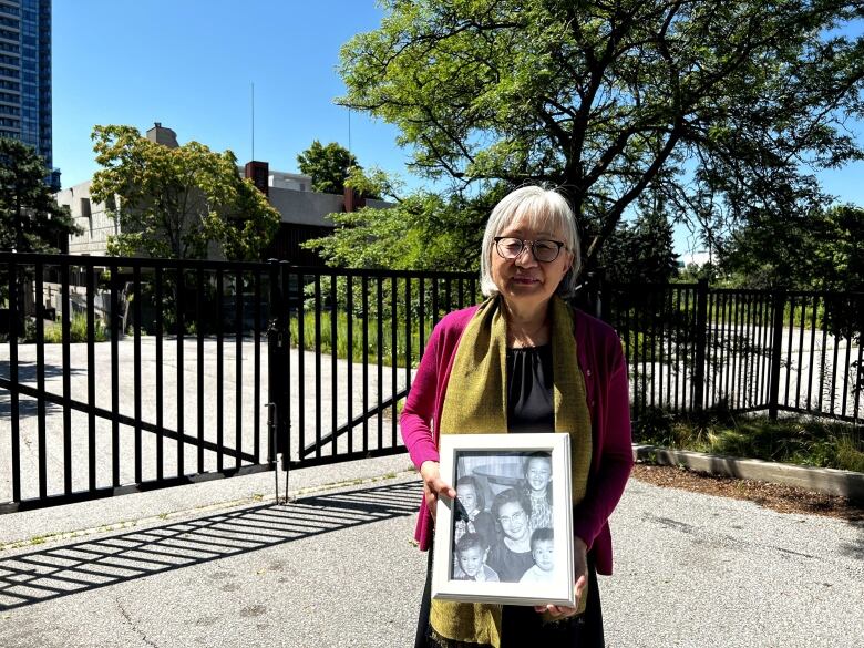 A senior Japanese woman holds a black and white photo in front of a shuttered community centre. 