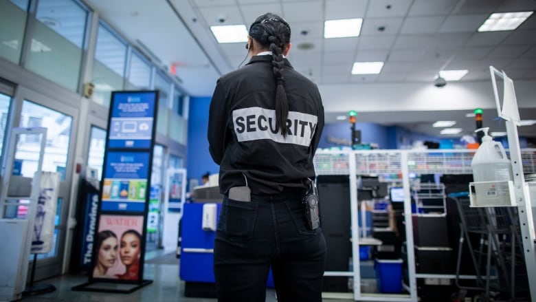 A female security guard with a long braid in her hair and the word Security on the back of her shirt is seen from behind. 