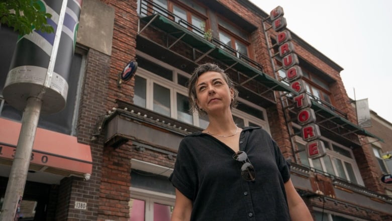 a woman with grey hair and a black tee shirt standing in front of the closed-down Drugstore bar in Montreal.