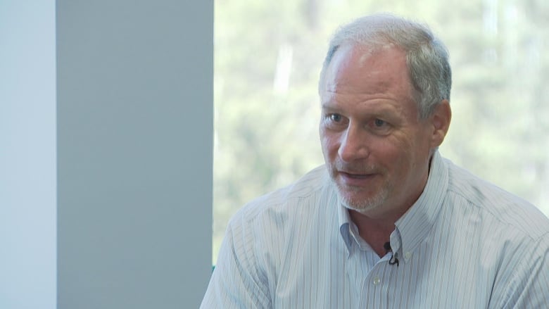 A man with grey hair and a beard wearing a collared shirt looks off to the side of the camera 