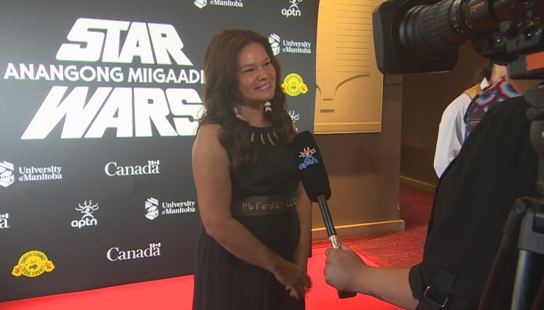 A woman stands on a red carpet, wearing a dress and talking to reporters.