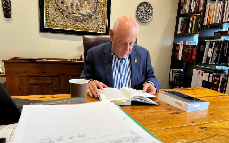 A bald man, wearing a blue blazer and blue and white striped shirt, sits at a desk reading a book.