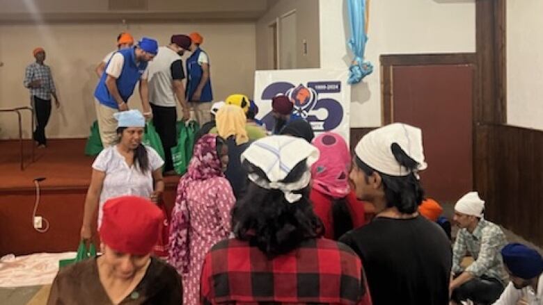 People line up in the Sikh temple while people on stage at the front distribute grocery bags of food. 