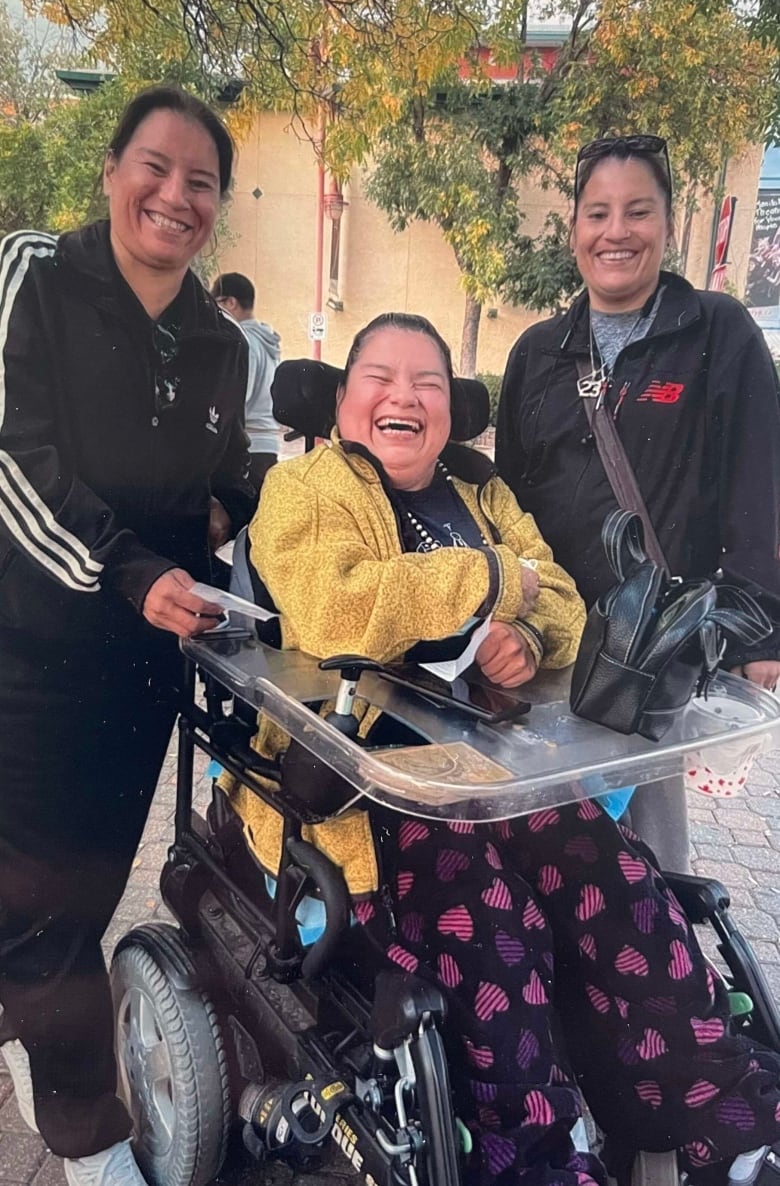 Three women, one of them in a wheelchair, stand in a park. 