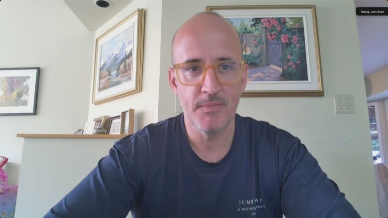 Man with glasses sits with flower painting in background.