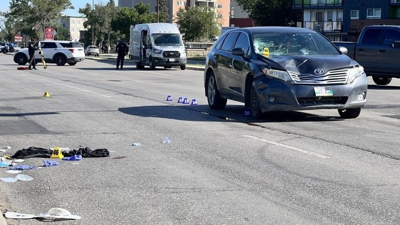 A damaged car is seen parked on a street, with personal items strewn on the road next to the vehicle.