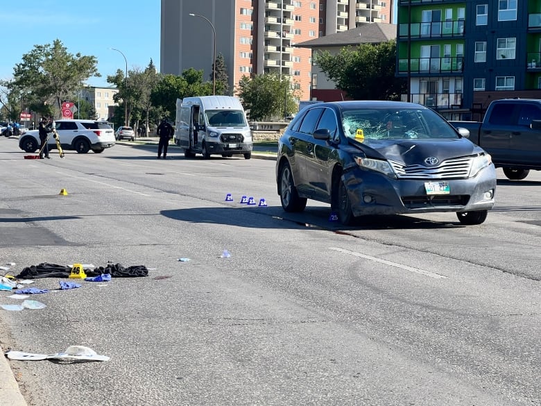 A damaged car is seen parked on a street, with personal items strewn on the road next to the vehicle.