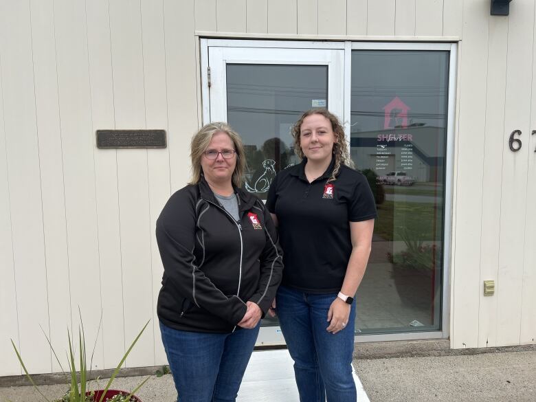 Two women in black tops stand in front of a glass door.