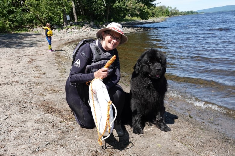 A woman sits by a river with her black dog. 