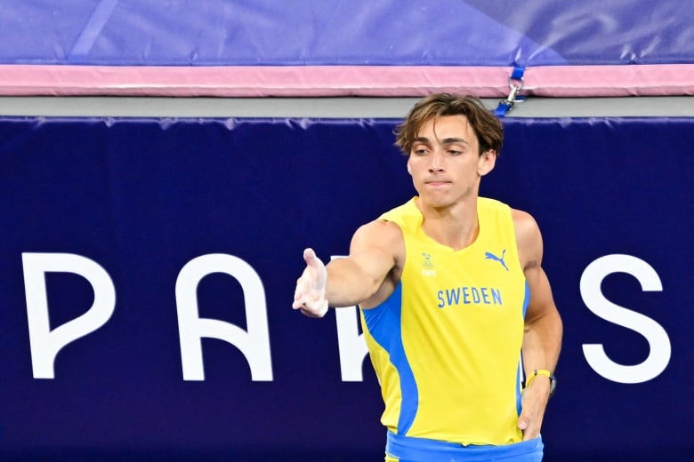 A male pole vaulter makes a gun shape with his outstretched right arm as he tucks his left hand into his shorts.