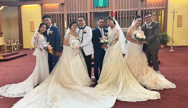 Four couples dressed in wedding attire. The women wearing bridal dressers and the men wearing suits. They are all standing in an alter inside a church. 