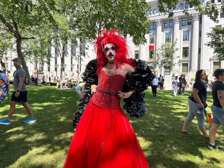 A person with fiery red hair, red and black makeup and wearing a corsetted red dress poses outdoors. 