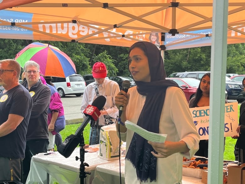 A woman in a head scarf stands under an orange tent outside in daylight with a microphone in her hand. She is surrounded by a small crowd