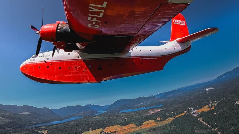 A red and white water bomber plane can be seen flying by with lakes, hills and forest below.