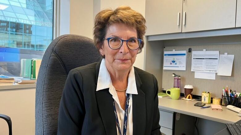 A woman wearing a suit sits at a desk and smiles into the camera.