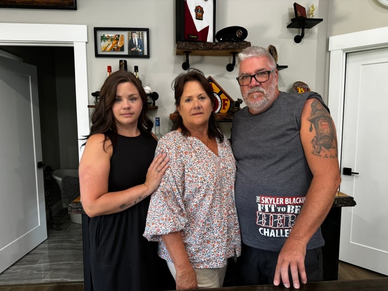 A woman with dark hair and wearing a black dress places a hand on a woman wearing a floral shirt. A man is standing with them with a firefighter tattoo.