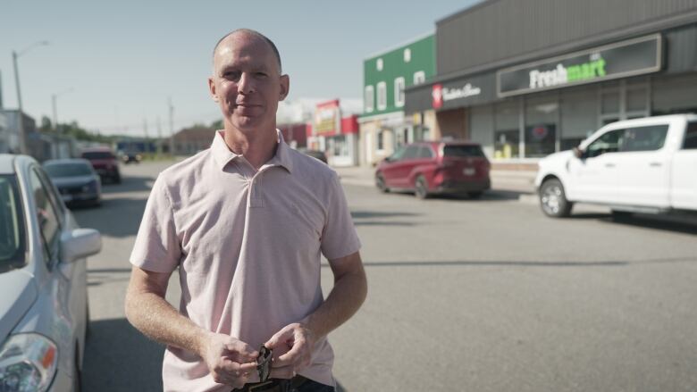 A bald white man wearing a baby pink collared short sleeve polo standing in the parking lot of a plaza