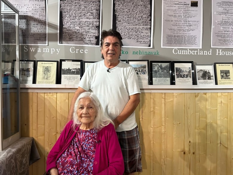 A man in a white shirt stands behind a woman in a purple sweater sitting in a wheelchair. Both are smiling for the camera.