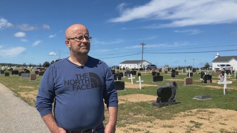 A bald man in a blue shirt with glasses on stands in front of a cemetery and a church.