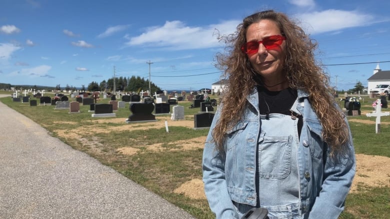 A woman with red sunglasses and a blue denim jacket and overalls stands in front of a cemetery. 