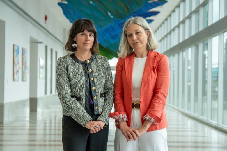 Two people wearing blazers stand beside each other in a hallway.