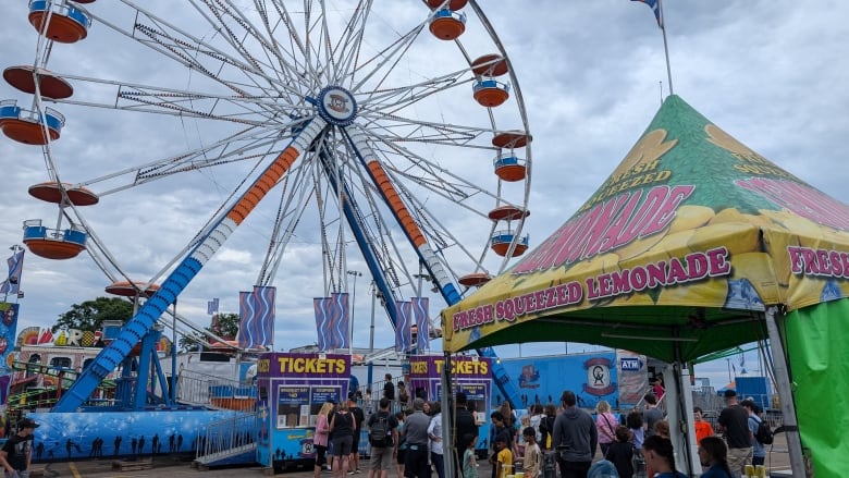 A ferris wheel, ticket booth and lemonade stand are seen against a cloudy sky.