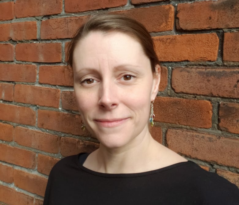 A smiling woman wearing a black dress stands in front of a brick wall.