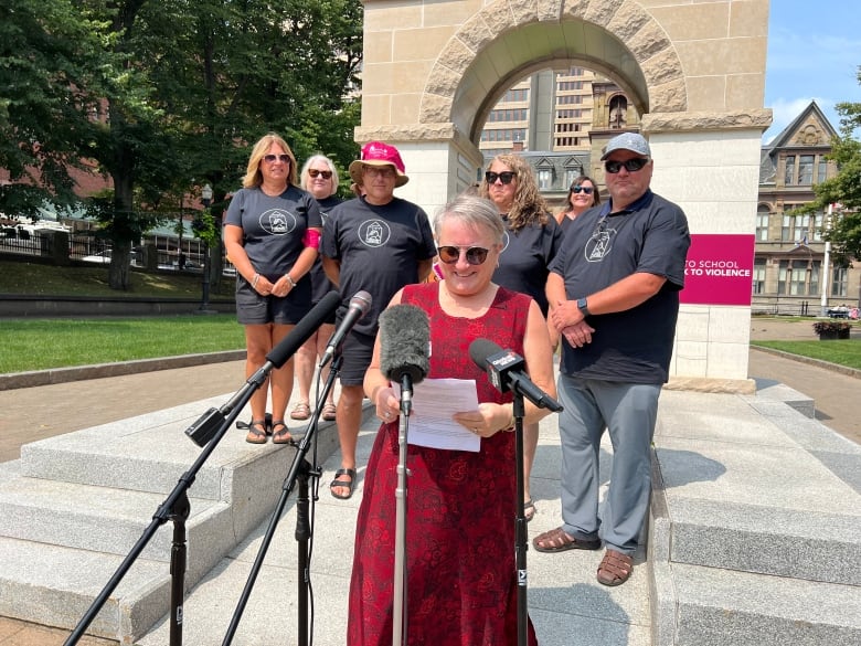 A group of people stand in front of a stone monument, with several microphones in front of them.