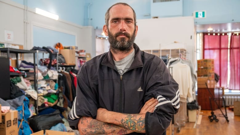 A person is seen standing in a room with their arms crossed. Behind them are racks and boxes of clothing.