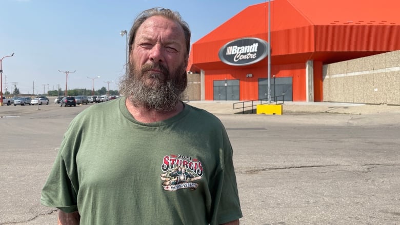 A man in a green shirt stands in a parking lot in front of an orange building. Over the man's right shoulder is a sign that reads 