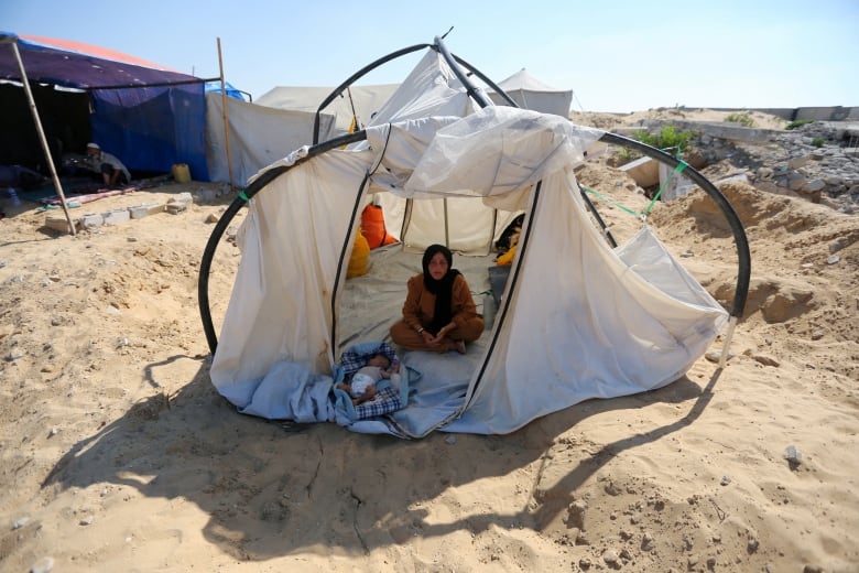 A woman sits on the floor of a white tent. A baby lies on the floor.