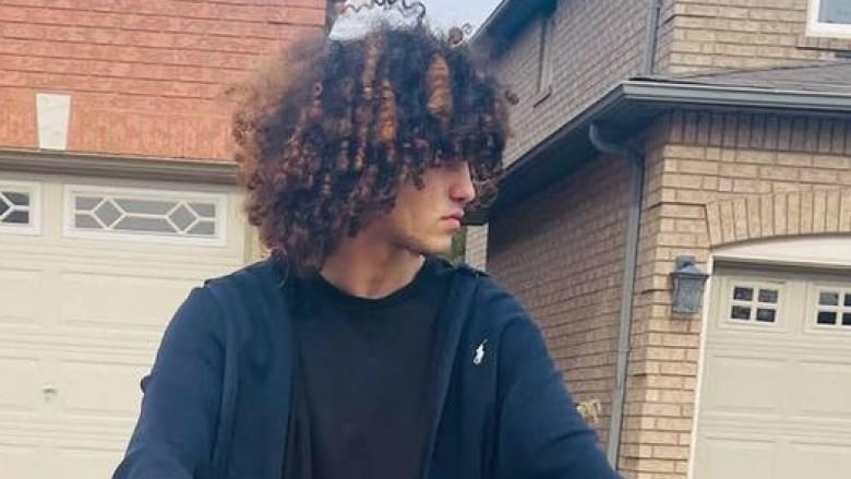 Man with long, curly hair on a bicycle in front of a brick house