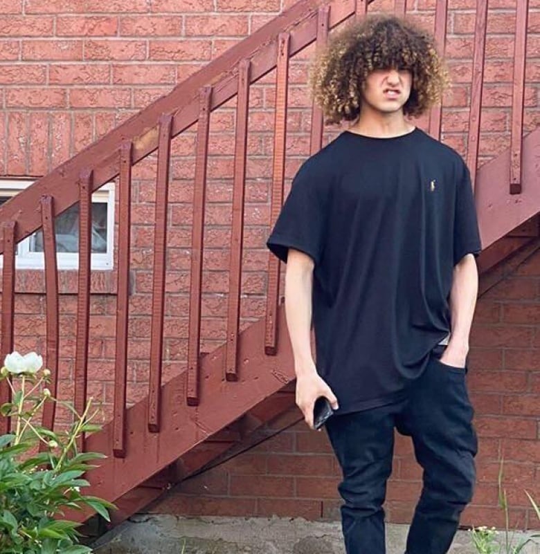 A man with long, curly hair stands in front of a brick wall.