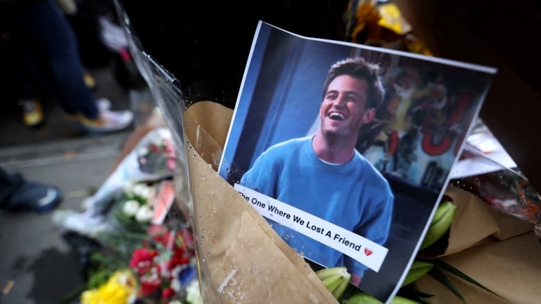 A photo of a  smiling man that says 'the one where we lost a friend.' flowers in the background