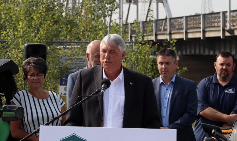 A man with grey hair in a suit jacket standing at a microphone with several other people behind him and a bridge and trees in the background.