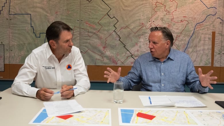 Two middle aged white men sit at a table with maps on it. 