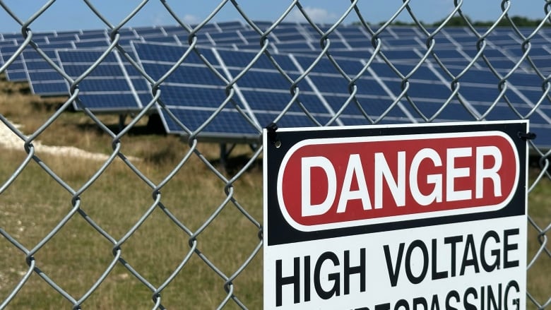 Solar panels are seen in the background behind a sign that says 'Danger High Voltage' on a chain-link fence.   