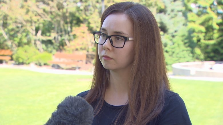 A woman with brown hair and glasses wearing a black shirt is interviewed in a park.