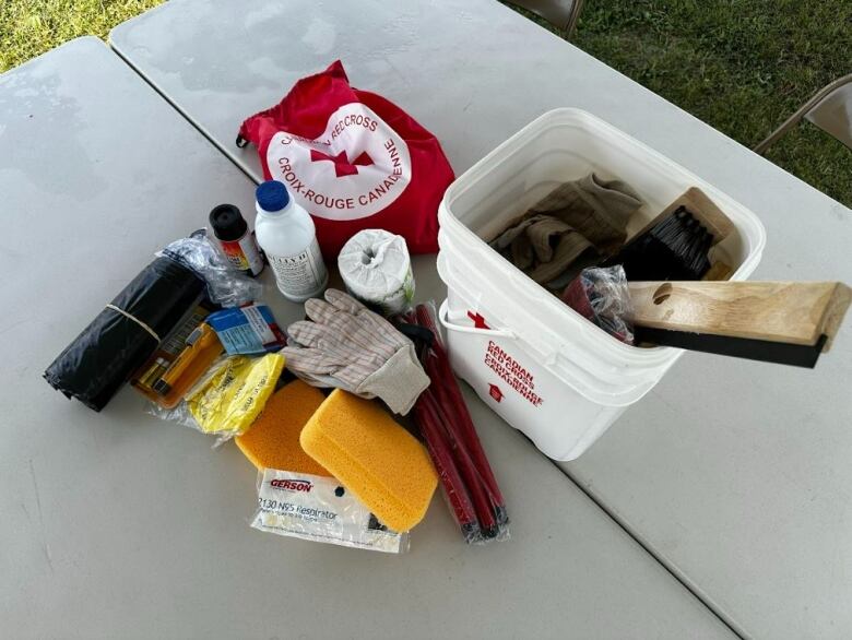 Multiple items are laid out on a foldable table. A white bucket with the Red Cross logo contains cleaning equipment; sponges and gloves are on the table, beside a red Red Cross bag.