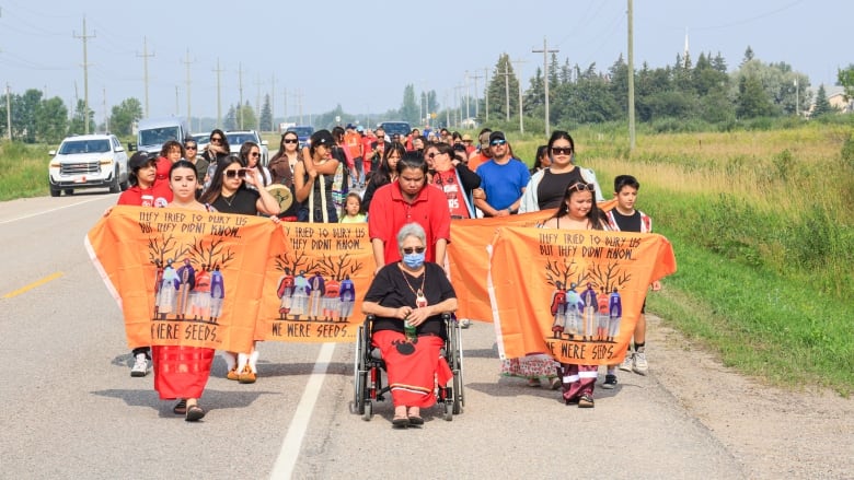 People, some of them holding orange banners and wearing red dresses, walk on a road.