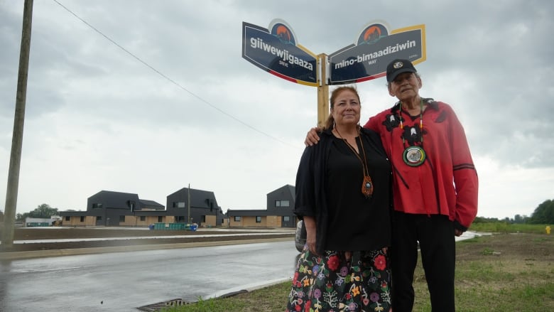 A man has his arm around a woman. Behind them are street signs and a community of houses.