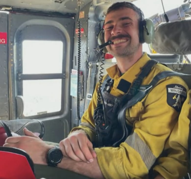 A firefighter with short brown hair and yellow jumpsuit smiles while taking a trip in a helicopter.