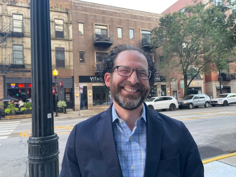 A man wearing a suit jacket and a plaid shirt smiles.