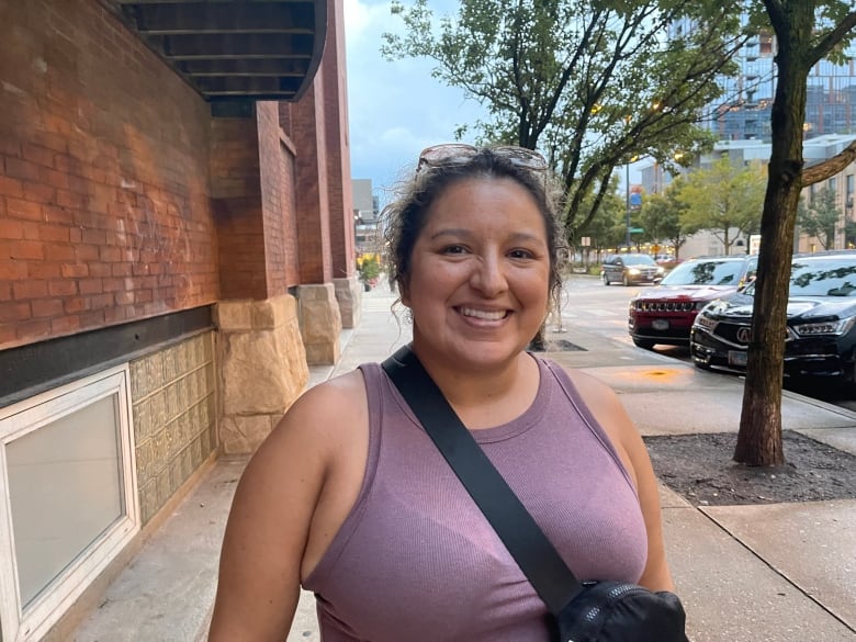 A woman wearing a crossbody bag and a purple tank top smiles.