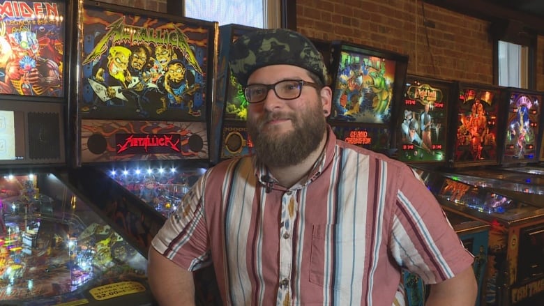 A man in a striped shirt and a hat sits in front of pinball machines.